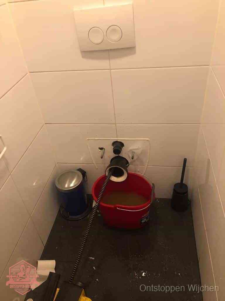 Een ontstopte wand-wc met verwijderde toiletpot waar een ontstoppingsveer in de wanduitgang verdwijnt, het achterhalen waar de verstopping zit in Wijchen