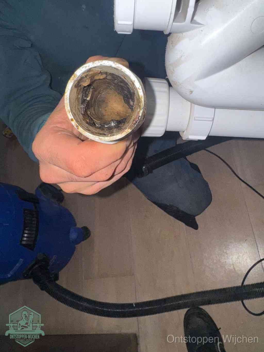 Een hand houdt een losgekoppeld PVC-afvoerbuisstuk vast dat van binnen volledig is dichtgeslibd met vet, vuil en aanslag, de meest voorkomende oorzaak van een verstopte afvoer in Wijchen