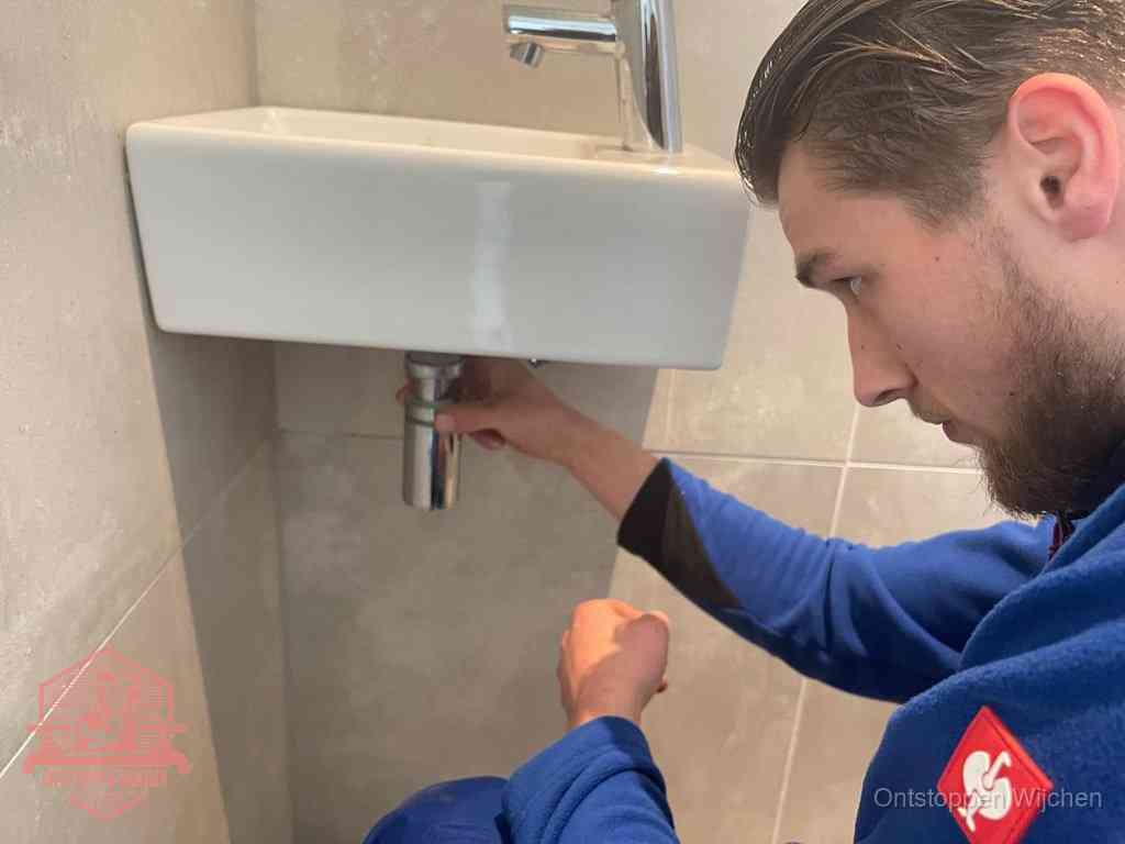 Een monteur draait met de hand de zwanenhalssifon onder een fonteinwasbak in de toiletruimte los om de bron van rioollucht in Wijchen te vinden