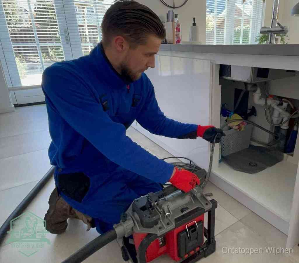 Een monteur met rode handschoenen knielt in een keuken en voert een elektrische ontstoppingsveer in tijdens het professioneel afvoer ontstoppen in Wijchen